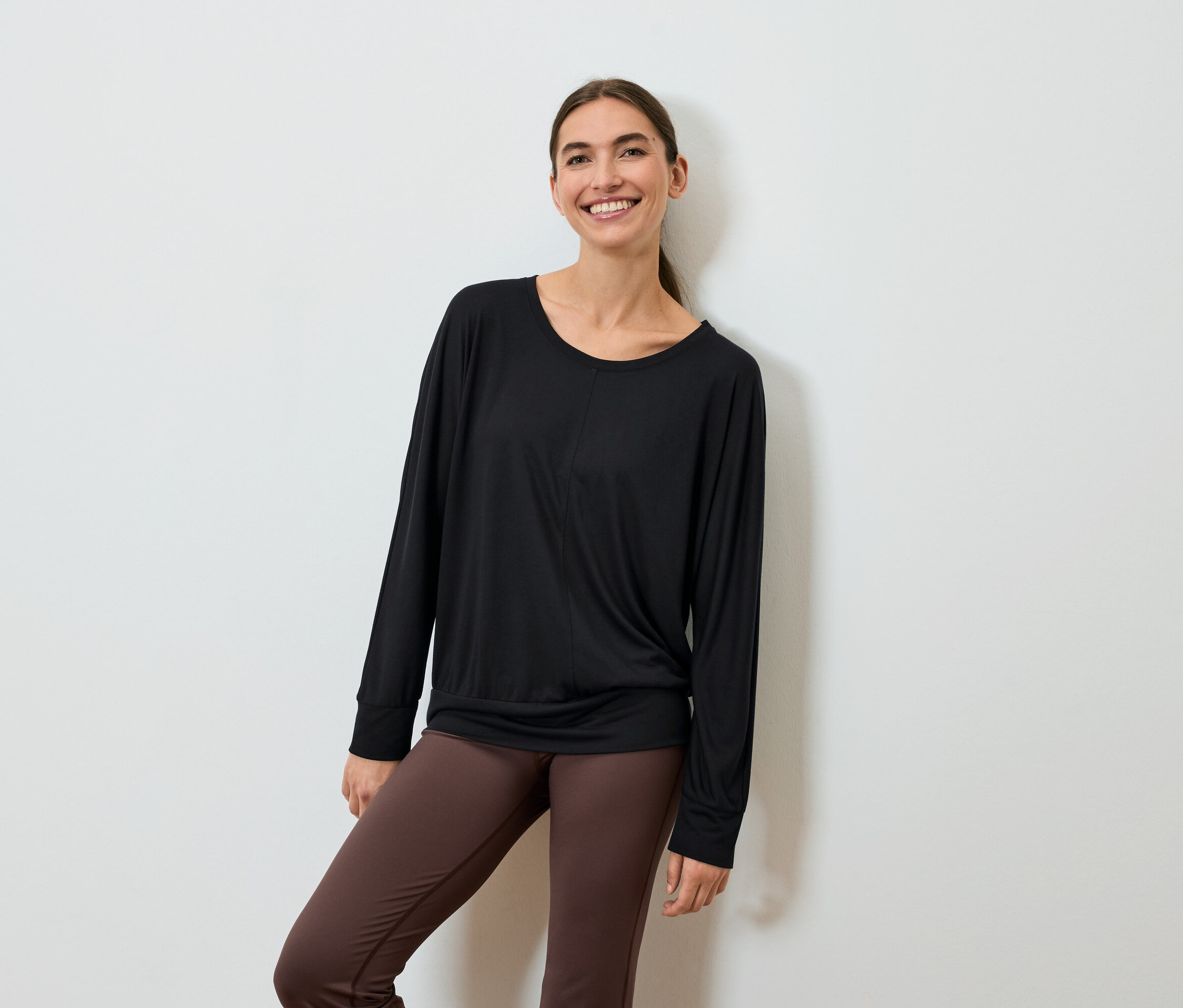 Lächelnde Frau in schwarzem Langarmshirt und braunen Leggings posiert an einer Wand.