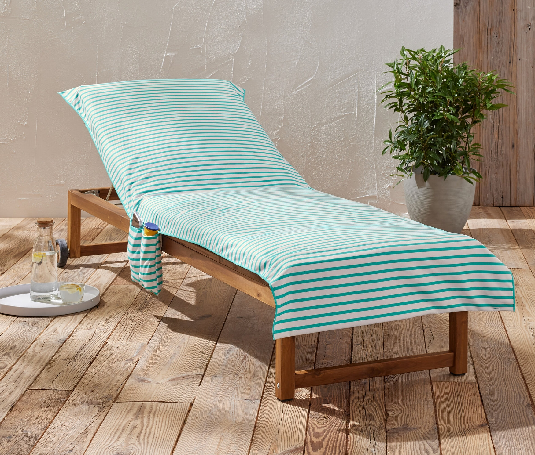 Gestreiftes Outdoor-Badetuch liegt auf einer hölzernen Sonnenliege neben einem Getränk und einer Pflanze.