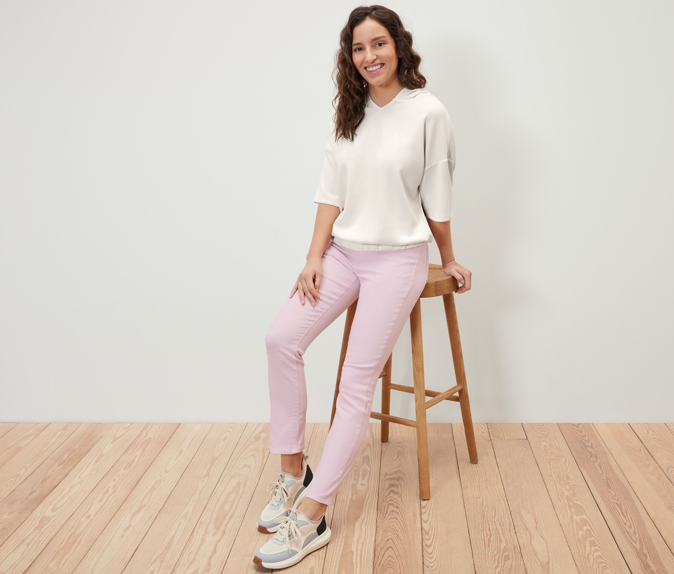 Frau sitzt auf einem Hocker und trägt einen cremeweißen Hoodie aus Modal-Mix und eine rosafarbene Hose.