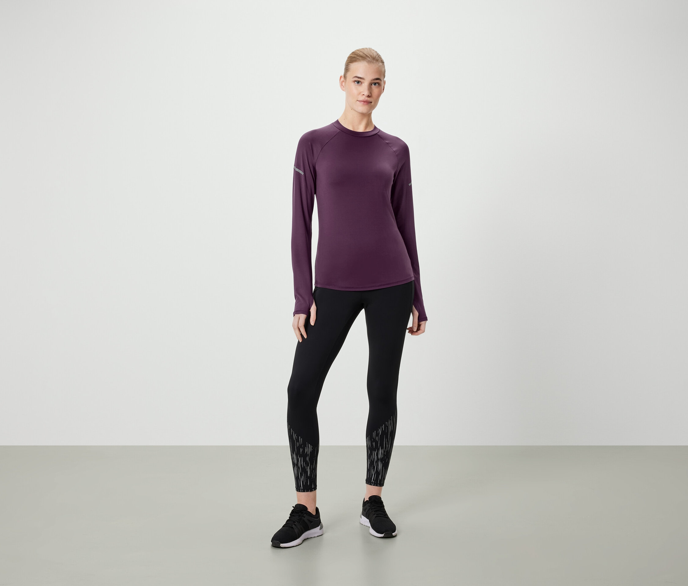 Frau steht in einem dunkelvioletten Langarmshirt, schwarzen Leggings mit Muster und schwarzen Schuhen.