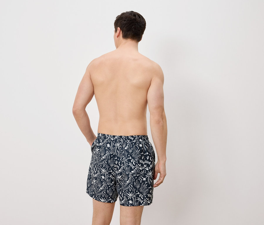 Mann von hinten, mit Badehose mit weißem Muster auf blauem Grund, die Hand in der Hosentasche.