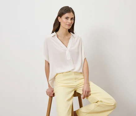Frau sitzt auf einem Hocker, trägt eine weiße Bluse und gelbe Hose.