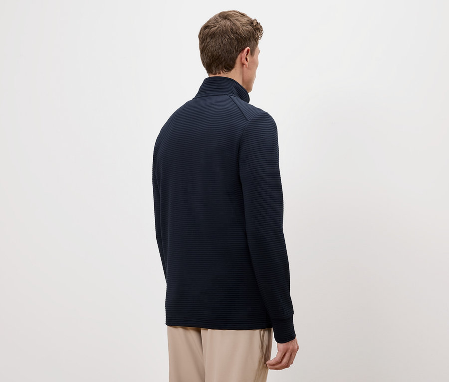 Rückansicht eines Mannes mit braunen Haaren, der einen dunkelblauen Pullover mit Stehkragen und beigefarbene Hosen trägt.