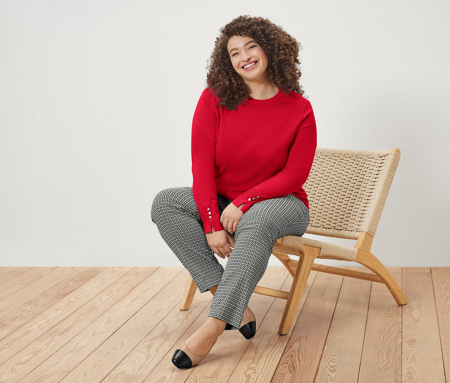 Eine Frau mit lockigem Haar sitzt lächelnd auf einem Stuhl und trägt einen roten Pullover und gemusterte Hosen.