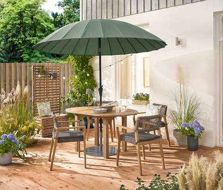 Ein runder Holztisch mit vier stapelbaren Gartenstühlen mit Textilgeflecht steht unter einem grünen Leco Shanghai Sonnenschirm. Eine Solar-Lichterkette »Glühbirnen« hängt über dem Tisch, auf dem eine LED-Akku-Outdoor-Tischleuchte, salbei steht. Zwei Outdoor-Kissen mit floralem Muster liegen auf einem Holzkasten.