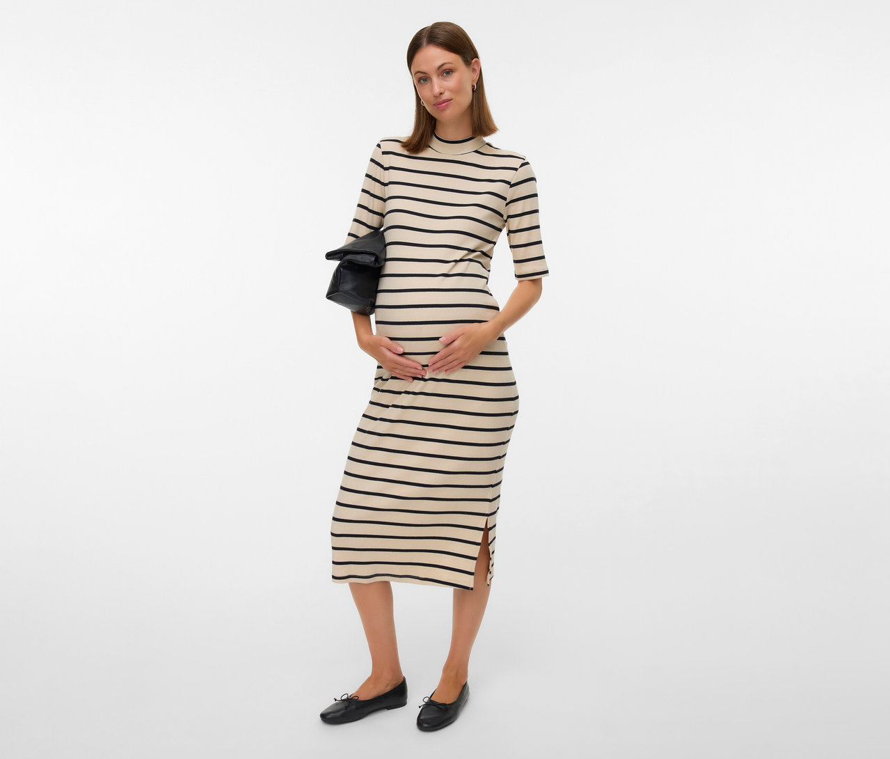 Schwangere Frau im MAMALICIOUS Kleid »MLLANE« mit schwarzen Streifen, hält eine schwarze Tasche und trägt flache, schwarze Schuhe.