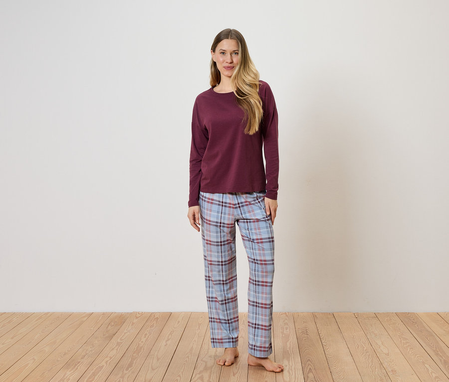 Eine Frau steht barfuß vor einer weißen Wand und trägt einen bordeauxroten Pullover und eine blau-karierte Pyjamahose.