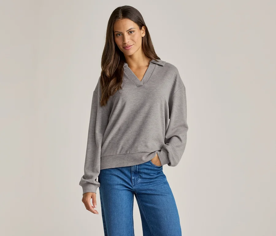 Frau mit braunen Haaren trägt einen grauen Pullover mit Kragen und blaue Jeans, eine Hand in der Hosentasche.