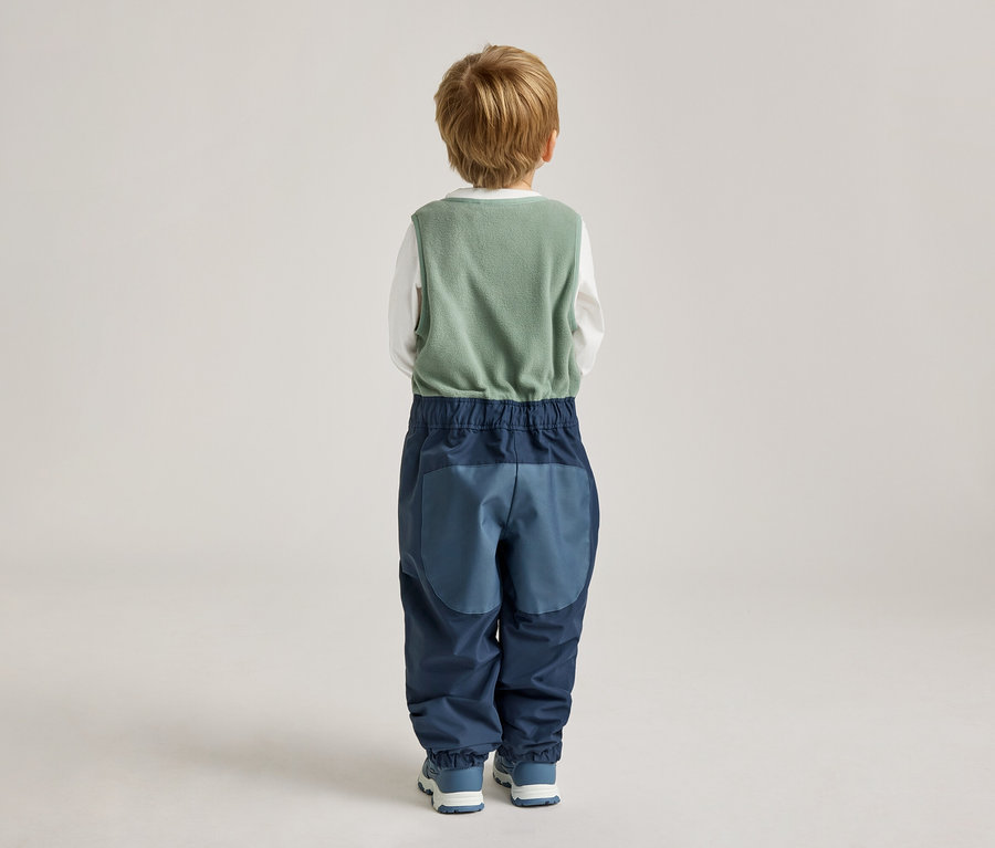 Rückansicht eines Kindes mit blonden Haaren, das eine grüne Weste, ein weißes Langarmshirt und blaue Hosen mit Farbblockierung trägt.