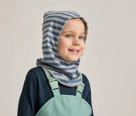 Lächelnder Junge mit blau-grau gestreifter Sturmhaube, marineblauem Langarmshirt und mintgrüner Latzhose vor hellgrauem Hintergrund.