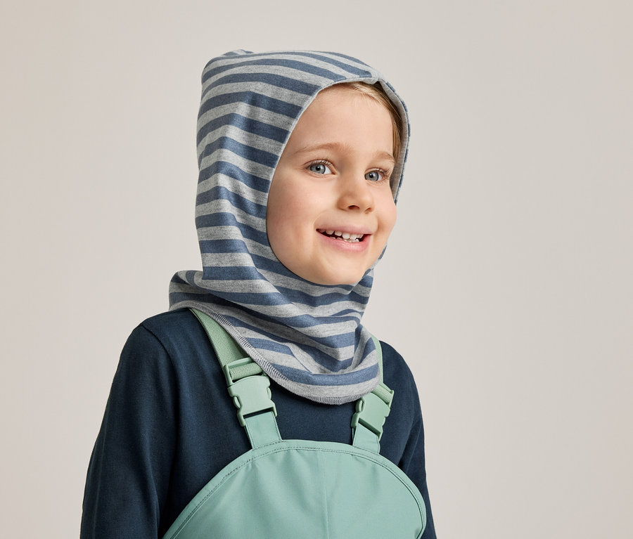 Lächelnder Junge mit blau-grau gestreifter Sturmhaube, marineblauem Langarmshirt und mintgrüner Latzhose vor hellgrauem Hintergrund.