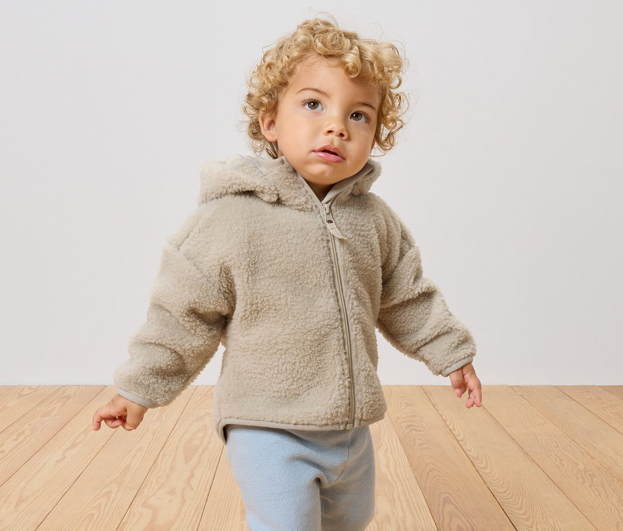 Ein Kleinkind mit blonden Locken trägt eine beige Teddyjacke mit Kapuze und hellblaue Hosen.