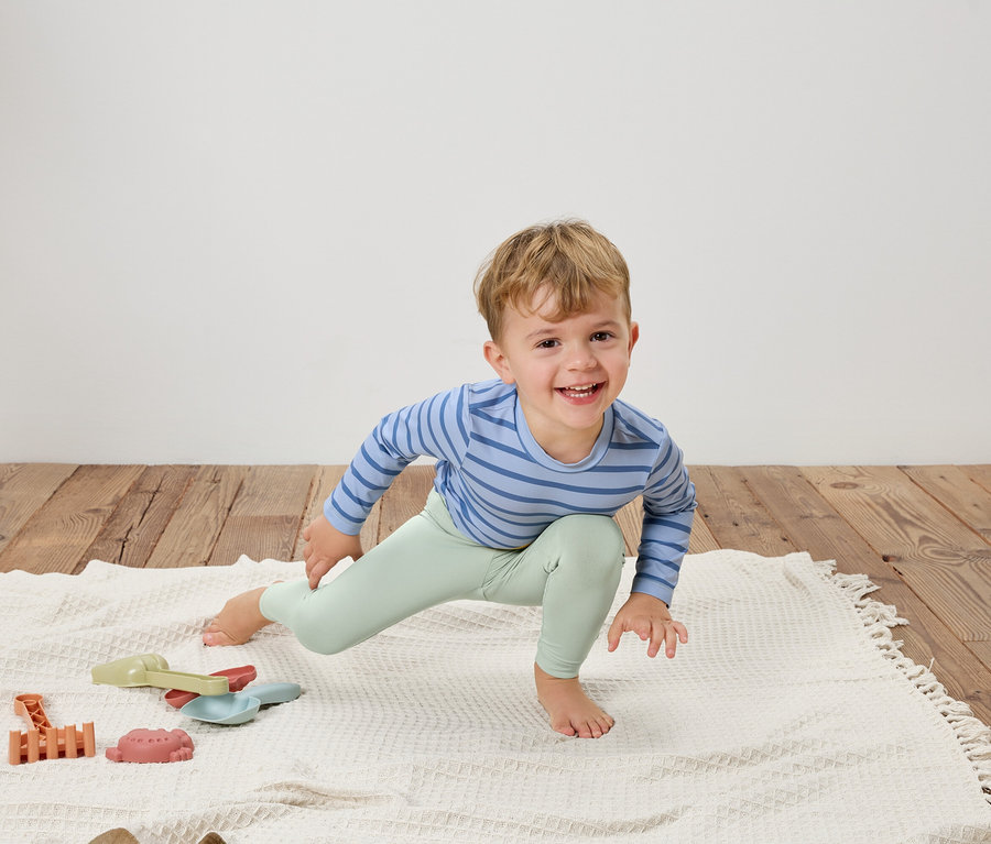 Ein lächelnder Junge in einem blau gestreiften Oberteil und hellgrünen Leggings dehnt sich auf einer weißen Decke neben Spielzeug.