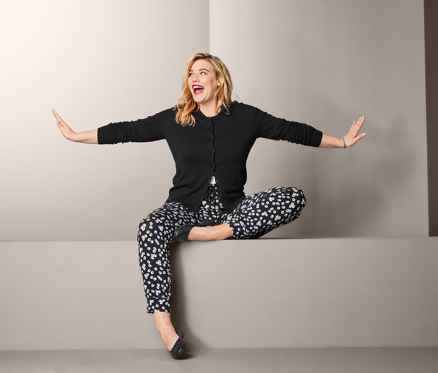 Blonde Frau sitzt mit ausgestreckten Armen und trägt eine Feinstrickjacke, eine Jerseyhose im Margeriten-Dessin, ein Halbarmshirt und Leder-Ballerinas.