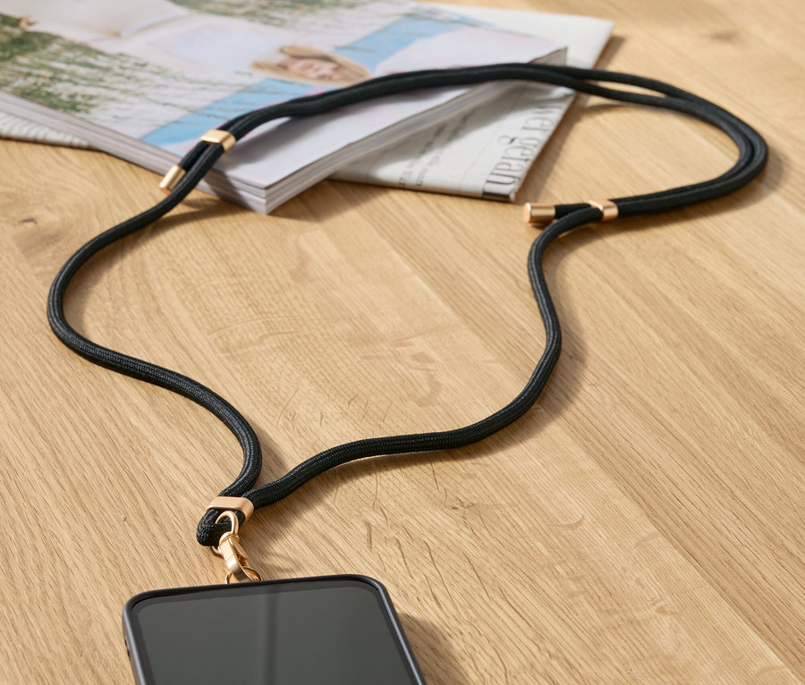 Schwarze Handykette mit goldenen Details liegt auf einem Tisch neben einer Zeitschrift und einer beigen Handtasche.