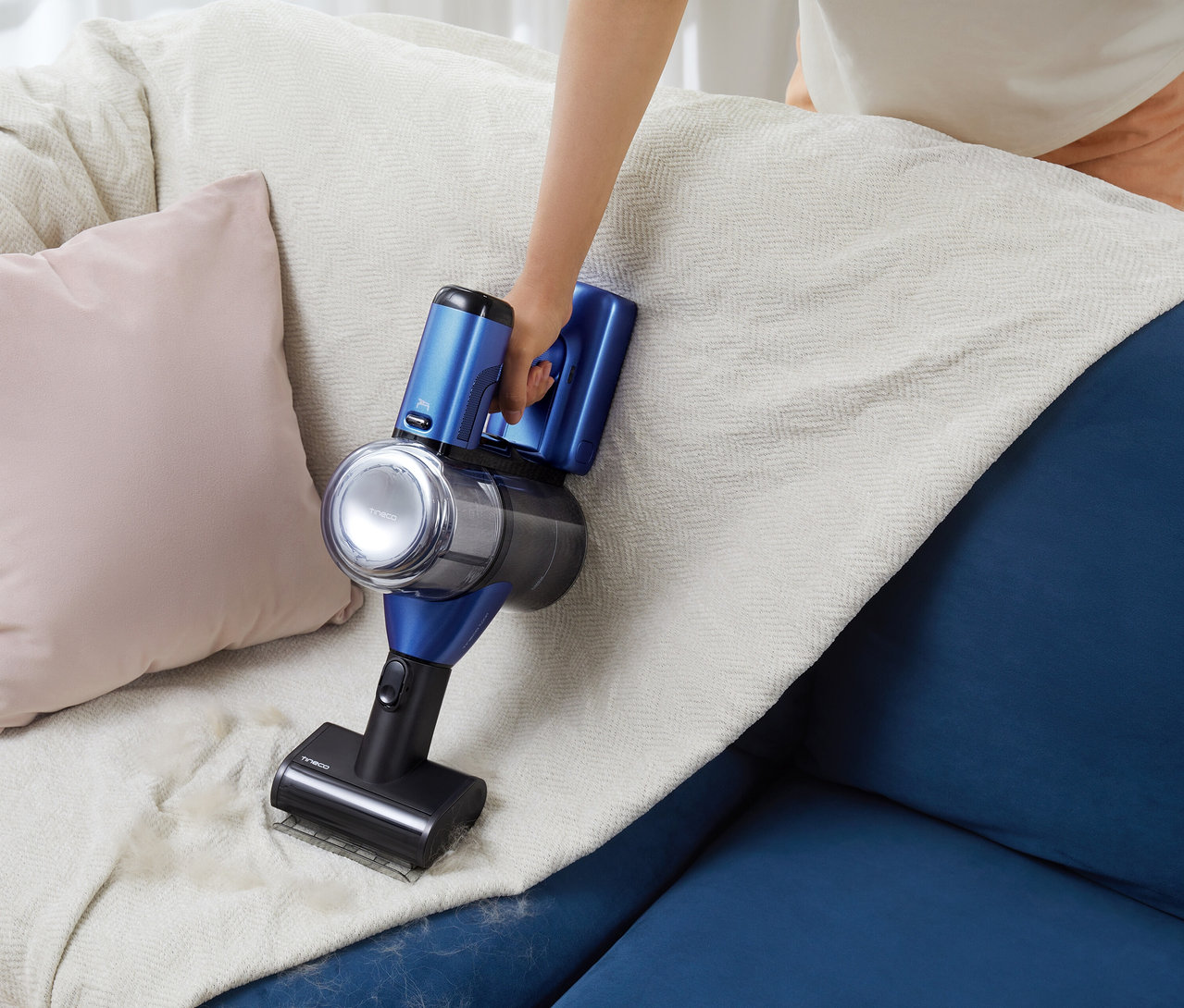 Eine Person verwendet einen blauen Tineco Akku-Staubsauger »Pure One S50 Pro«, um eine weiße Decke auf einem blauen Sofa zu reinigen.