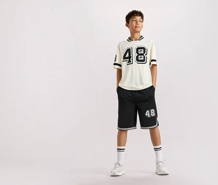 Ein Teenager steht mit Händen in den Hosentaschen, bekleidet mit einem beigen T-Shirt mit der Zahl 48, schwarzen Shorts mit der Zahl 48, weißen Socken mit Streifen und weißen Turnschuhen vor einem schlichten Hintergrund.