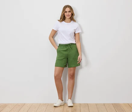 Frau in weißem T-Shirt und grünen Shorts mit Kordelzug, stehend vor einer weißen Wand.