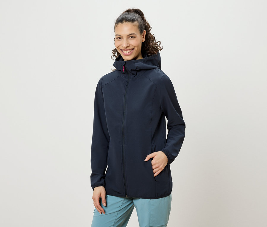 Frau mit hochgesteckten lockigen Haaren, die eine marineblaue Jacke mit Kapuze und blaue Hosen trägt und lächelt, wobei eine Hand in der Jackentasche steckt.