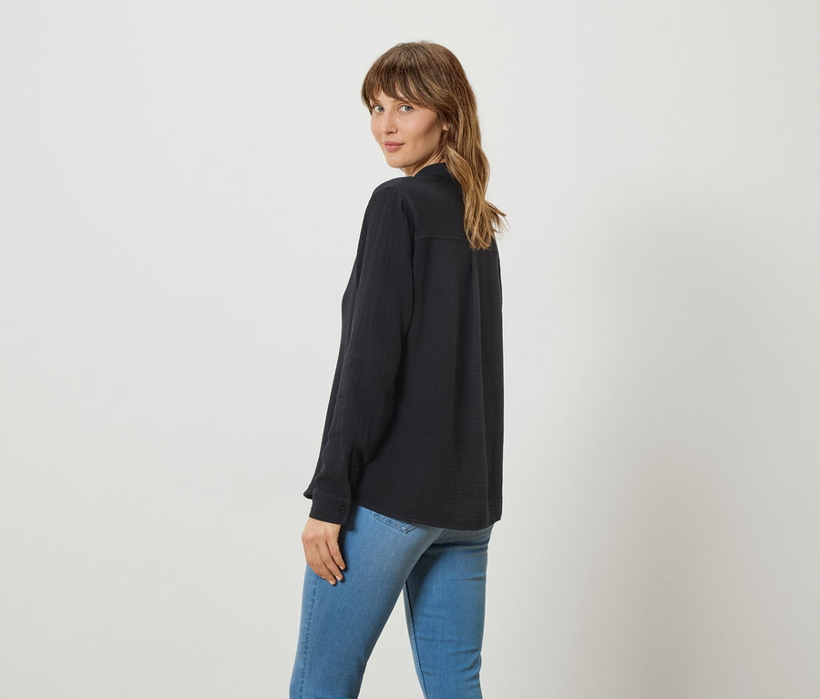 Frau mit langen gewellten Haaren, die eine lange, dunkle Bluse und Jeans trägt und über die Schulter in die Kamera blickt.