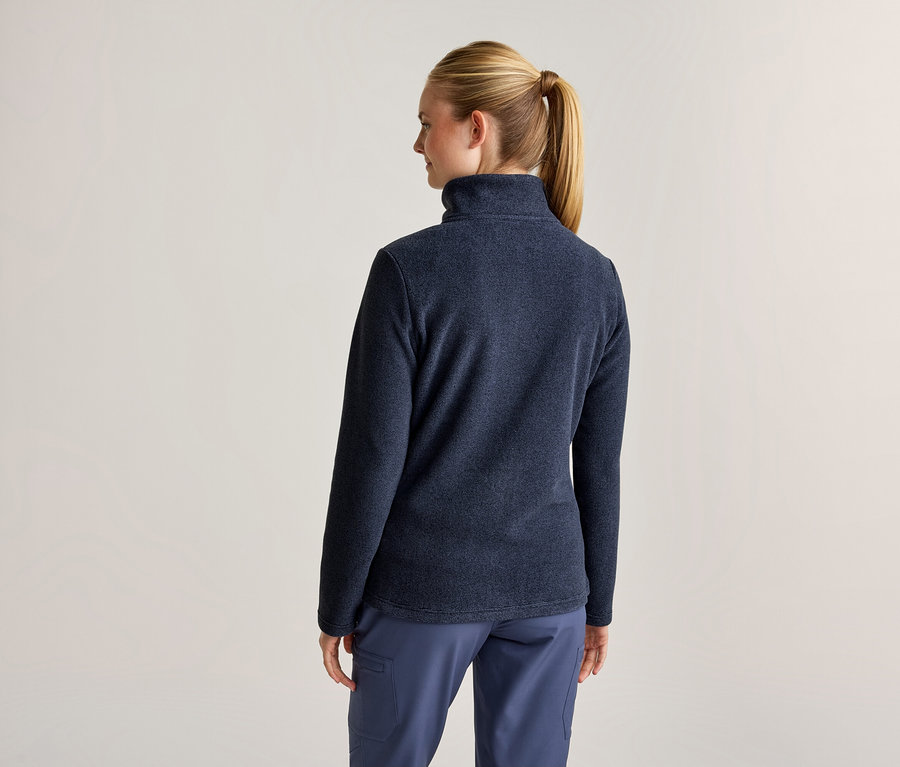 Eine Frau von hinten, die eine dunkelblaue Fleecejacke und eine blaue Hose trägt. Ihr blondes Haar ist zu einem Pferdeschwanz gebunden.