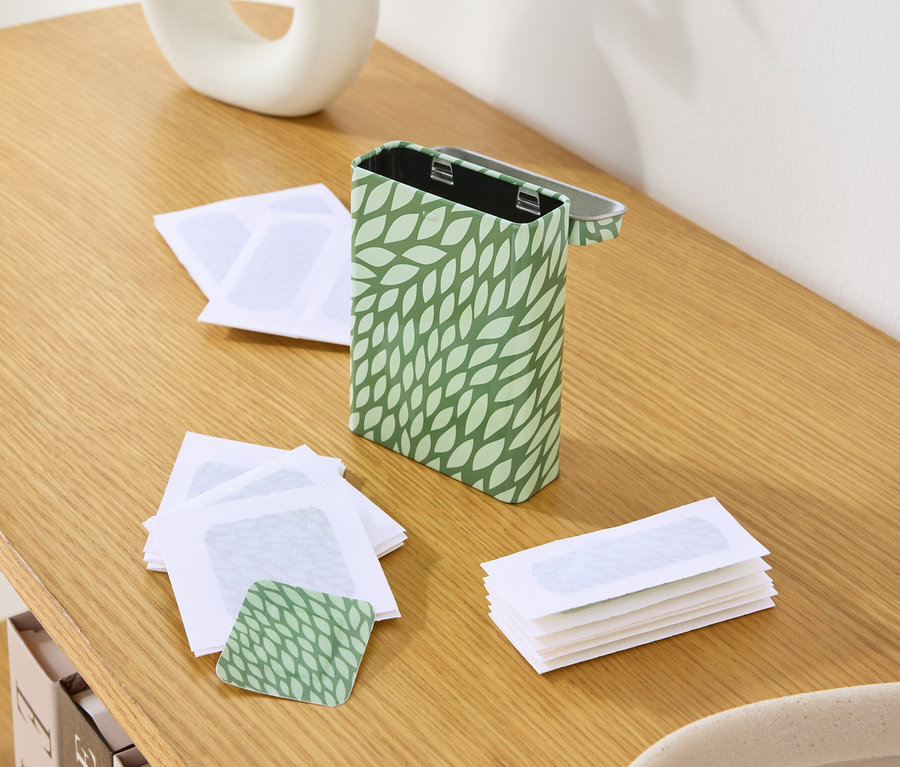 Auf einem Holztisch stehen eine Dose mit grünem Blattmuster, leere Wachsstreifen und eine Vase mit Pflanzen.