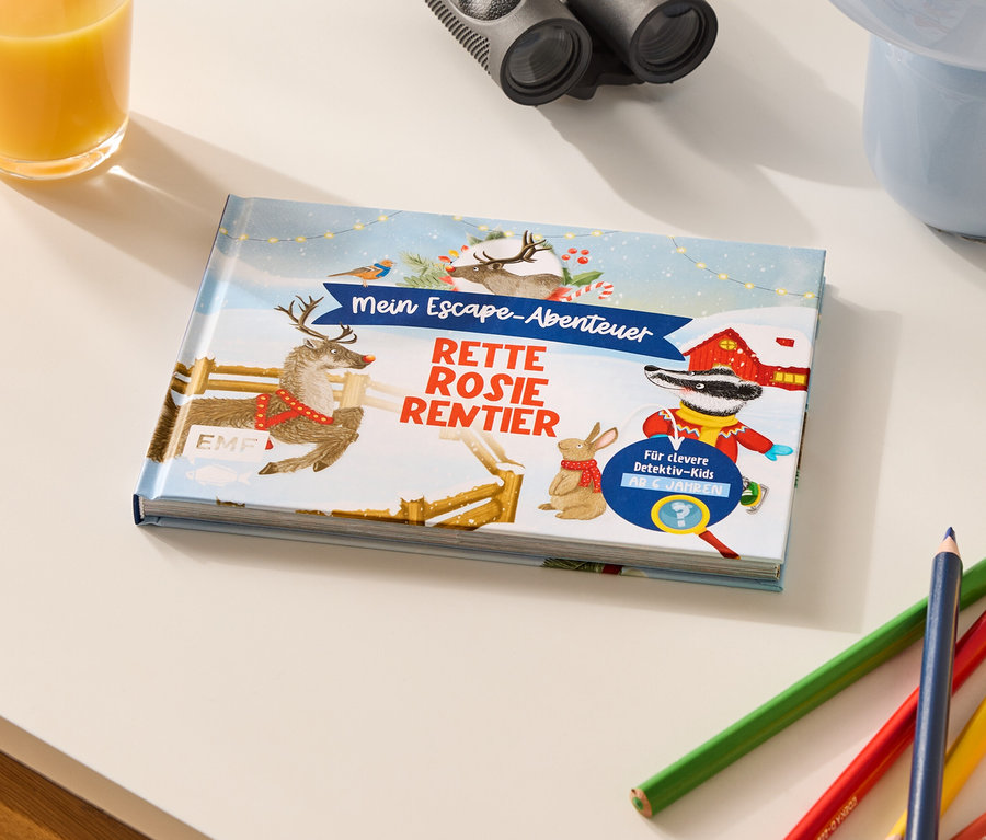 Ein Kinderbuch mit dem Titel "Mein Escape-Abenteuer: Rette Rosie Rentier" liegt auf einem Tisch, zusammen mit Buntstiften, einem Glas Orangensaft und einem Fernglas.