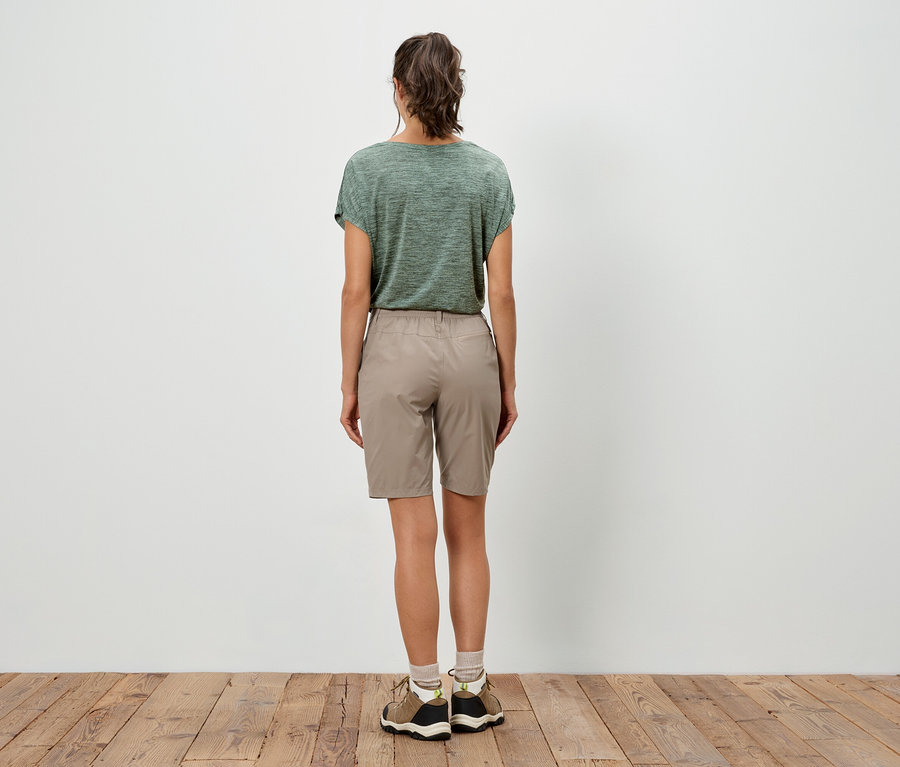 Eine Frau steht mit dem Rücken zum Betrachter, sie trägt ein grünes T-Shirt und beigefarbene Shorts.