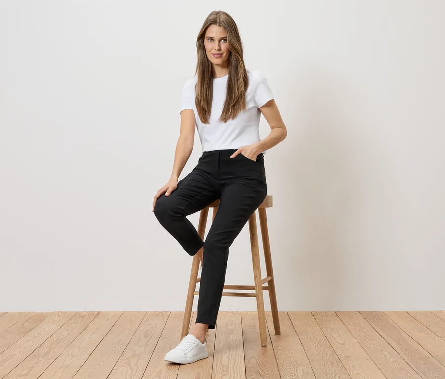 Frau mit weißem T-Shirt und schwarzen Hosen sitzt auf einem Hocker vor einer weißen Wand und einem Holzboden.