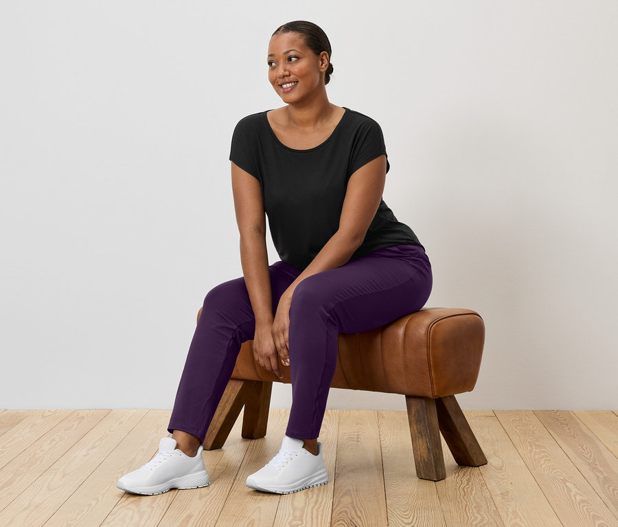 Eine Frau mit dunklen Haaren sitzt auf einem Turnbock und trägt ein schwarzes T-Shirt, eine violette Hose und weiße Turnschuhe.