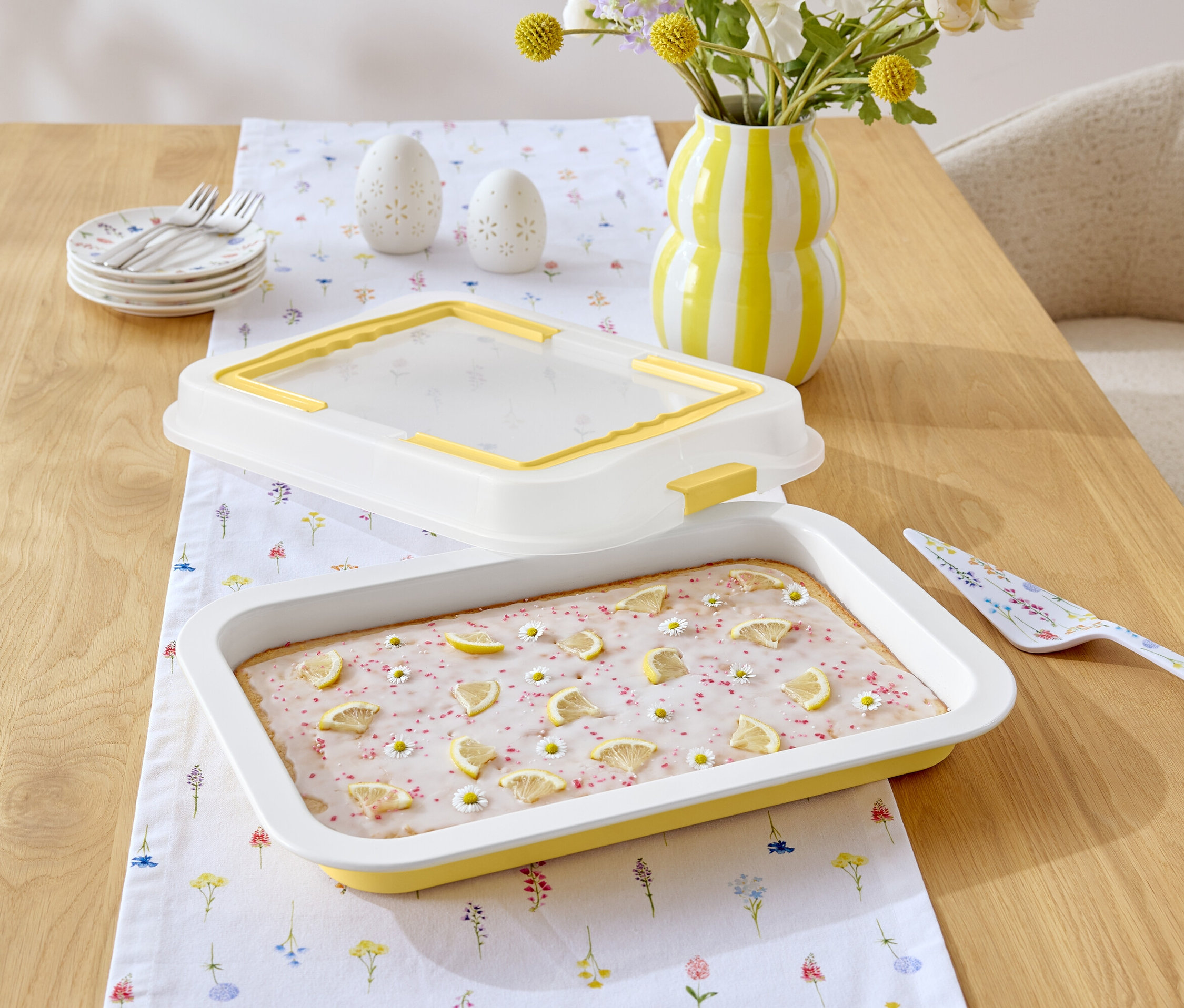 Auf einem Holztisch liegen ein Kuchen mit Zitronenglasur und Gänseblümchen, ein gelb-weiß gestreifter Krug, eine Tortenschaufel mit Blumenmuster und ein weißes Tischtuch mit Blumenmuster.