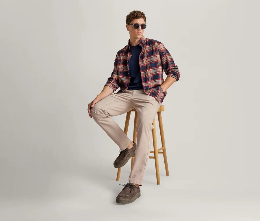 Junger Mann mit Sonnenbrille, kariertem Hemd, marineblauem T-Shirt und beiger Hose sitzt auf einem Holzhocker.