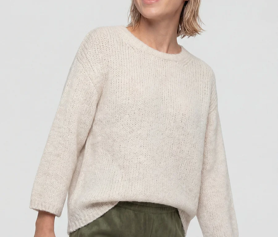 Eine lächelnde Frau mit kurzen blonden Haaren trägt einen hellen, gestrickten Olsen 3/4-Pullover und eine grüne Hose, blickt nach rechts.