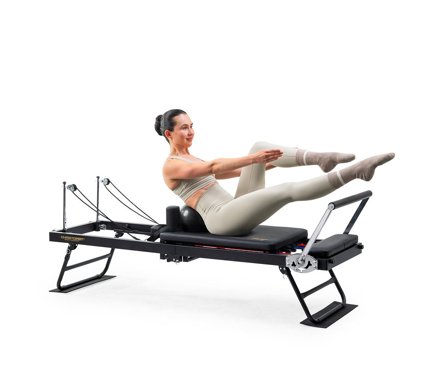 Frau in heller Sportkleidung trainiert auf einem schwarzen Pilates-Reformer, lehnt sich auf einen Ball zurück, Beine angehoben, Arme ausgestreckt.