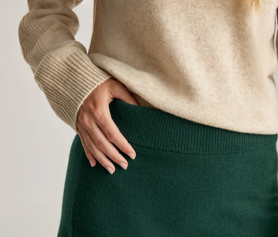 Nahaufnahme einer Person, die einen beigefarbenen Pullover mit Rippbündchen und einen dunkelgrünen Rock mit geripptem Bund trägt. Eine Hand ruht auf dem Rockbund.