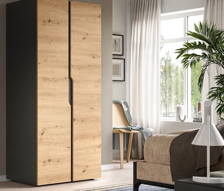 Modernes Schlafzimmer mit einem zweitürigen Kleiderschrank, links dunkelgrau und vorne helles Holzdekor. Ein gestreifter Teppich liegt am Boden, und ein Fenster mit weißen Vorhängen ist im Hintergrund, neben einem Holzstuhl mit einer Decke.
