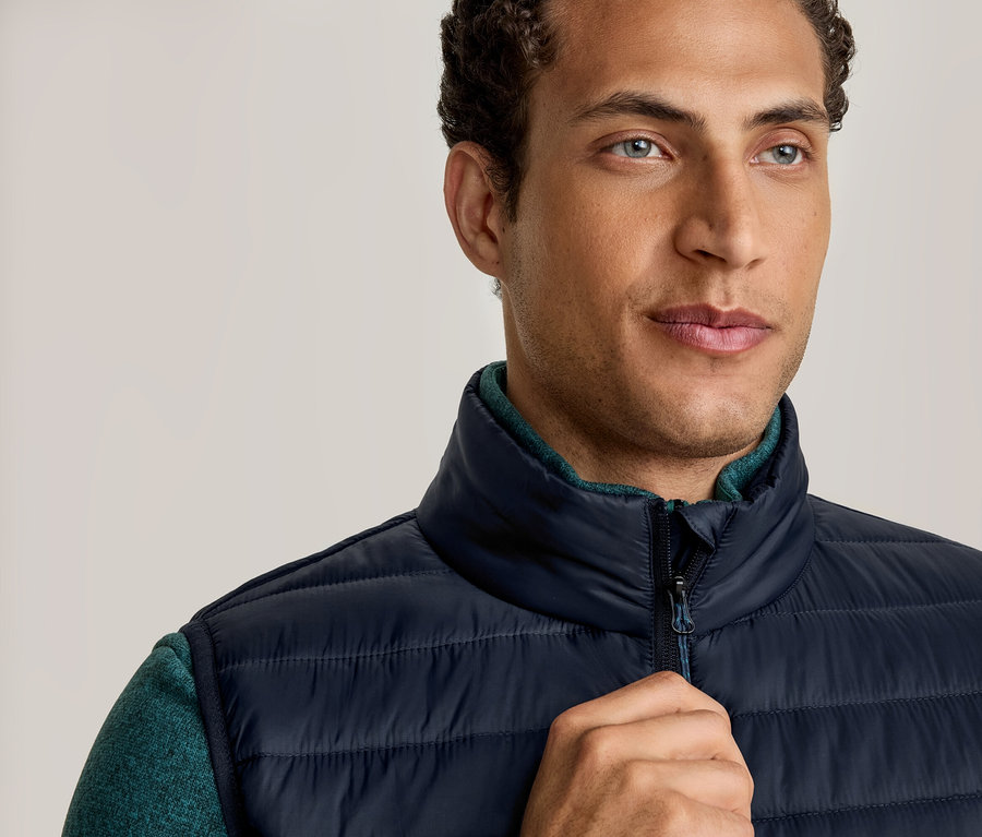 Ein Mann trägt eine dunkelblaue Steppweste über einem grünen Langarmshirt und hält seine rechte Hand am Reißverschluss der Weste. Er blickt leicht nach rechts.