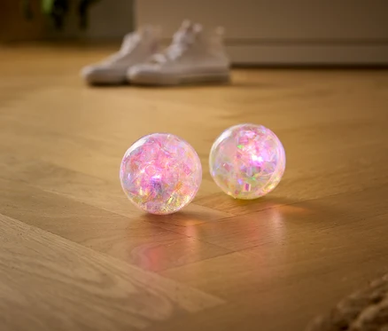 Zwei durchsichtige, schimmernde Glitzerbälle liegen auf einem Holzboden, im Hintergrund unscharf ein Paar weiße Turnschuhe.