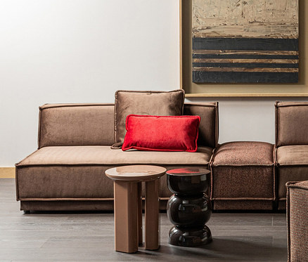 Ein braunes Modulsofa mit einem roten, rechteckigen Kissen steht auf einem dunklen Holzboden. Davor stehen zwei Beistelltische: ein hellbrauner mit drei Beinen und ein dunkler, sanduhrförmiger. Über dem Sofa hängt ein abstraktes Gemälde.