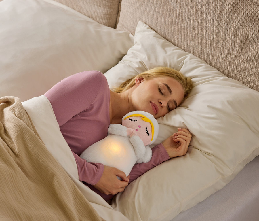 Eine blonde Frau schläft auf der Seite im Bett, trägt ein rosa Oberteil und kuschelt ein weißes Plüschtier mit leuchtendem Bauch unter einer beigen Decke.