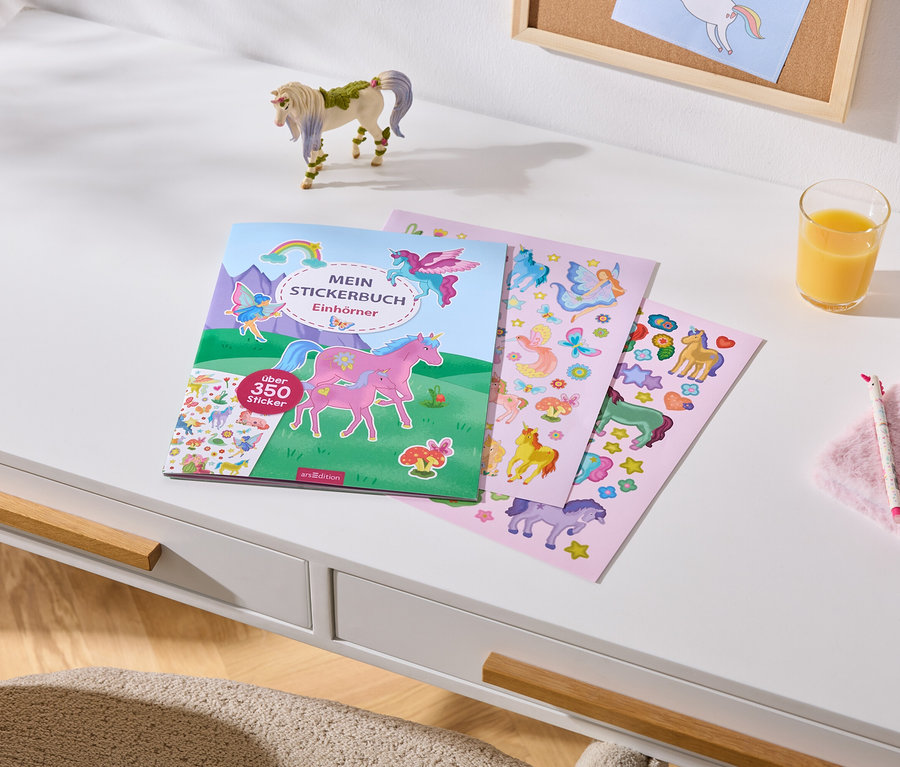 Weißer Schreibtisch mit einem Stickerbuch „MEIN STICKERBUCH Einhörner“ (über 350 Sticker), drei Stickerbögen, einer Einhornfigur, einem Glas Orangensaft, einem rosa Plüsch-Notizbuch, einem Einhornstift und einem Einhornarmband.