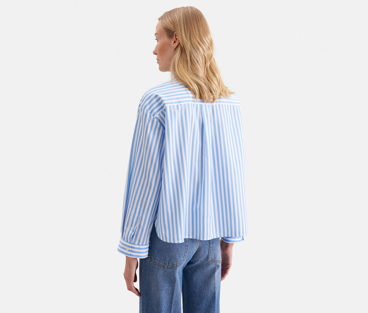 Frau mit blonden Haaren, von hinten gesehen, trägt eine blau-weiß gestreifte Seidensticker Oversize Hemdbluse mit langen Ärmeln und blaue Jeans.
