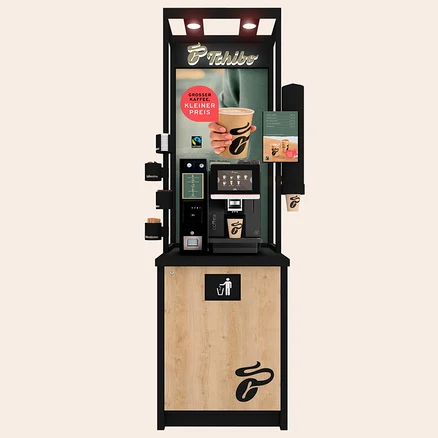 Eine schwarze und holzfarbene Tchibo Selbstbedienungs-Kaffeemaschine mit einem Bildschirm, der Kaffeeoptionen und eine Hand mit einem Kaffeebecher zeigt. Unten ist ein Mülleimer-Symbol.