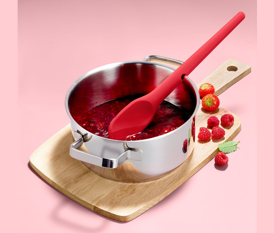 Ein Funktions-Kochlöffel liegt in einem Edelstahltopf mit Marmelade, der auf einem Holzbrett steht. Um ihn herum liegen Himbeeren.