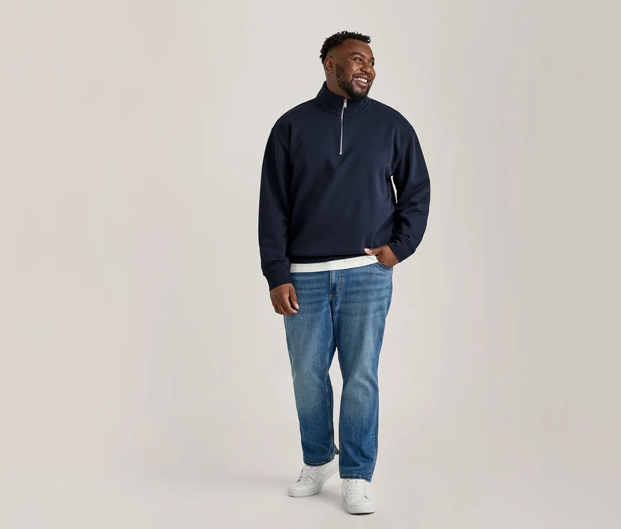 Ein lächelnder schwarzer Mann steht da, blickt nach rechts und trägt ein dunkelblaues Quarter-Zip-Sweatshirt, hellblaue Jeans und weiße Turnschuhe, eine Hand in der Tasche.