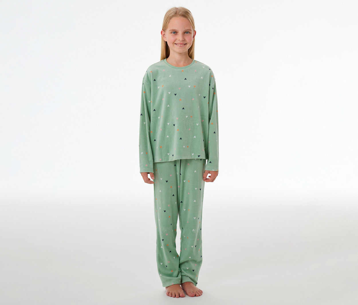 Lächelndes Mädchen steht barfuß in hellgrünem SCHIESSER Kinder-Velours-Pyjama »Herzchen« mit bunten Herzchenmustern.