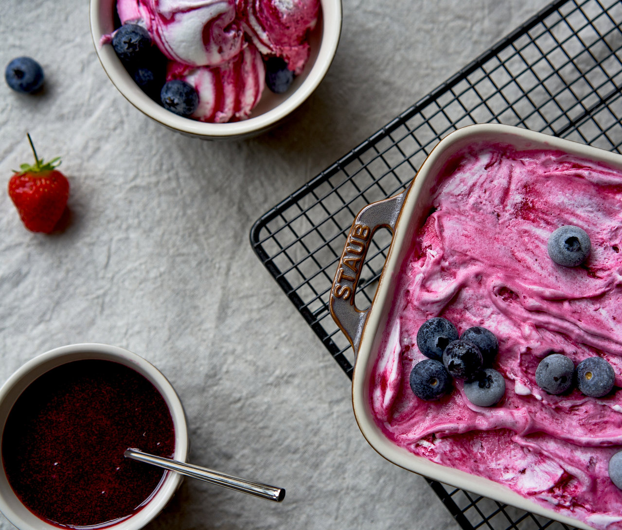 Blick von oben auf eine rechteckige, antikgraue STAUB Auflaufform mit pinkfarbenem gefrorenem Dessert und Heidelbeeren, daneben eine Schale mit Beeren-Eis, eine Schale mit Beerensauce und Löffel sowie eine Erdbeere auf hellgrauem Untergrund.
