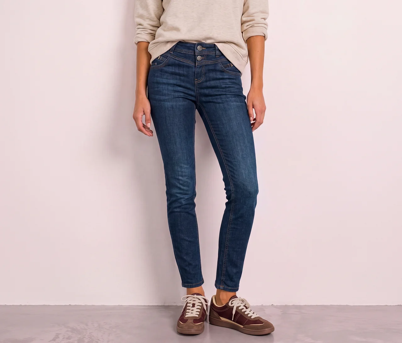 Eine Person steht und trägt ein beiges Langarmshirt, eine dunkelblaue hochgeschnittene Street One Jeans mit zwei Knöpfen und braun-beige Sneaker.