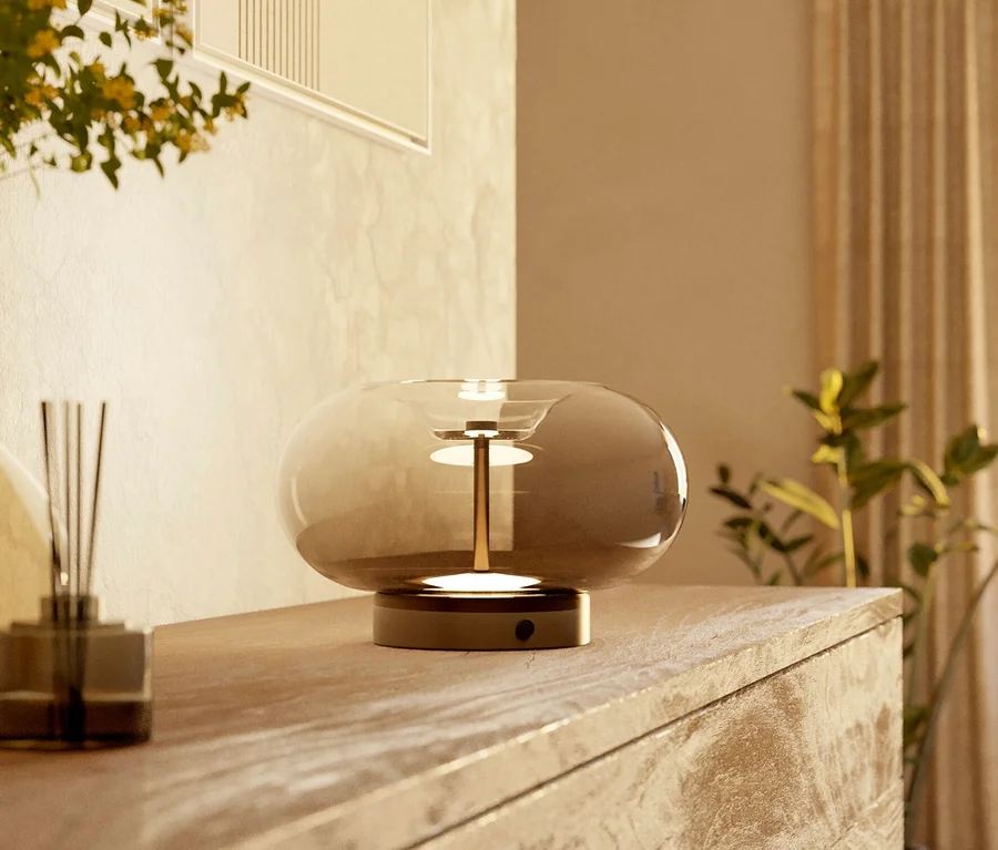 Eine moderne Tischlampe mit rauchfarbenem Glasschirm und goldenem Sockel beleuchtet eine helle Holzkommode neben einem dunklen Glasdiffusor. Im weich beleuchteten Hintergrund sind ein gerahmtes abstraktes Kunstwerk und Pflanzen zu sehen.