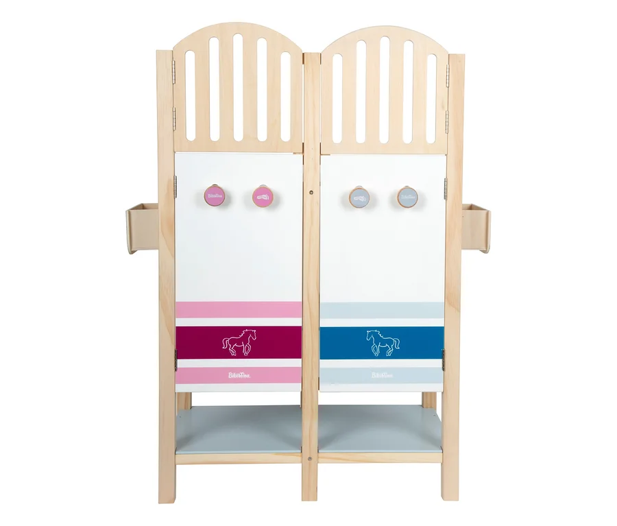 Ein hölzerner small foot Pferdestall »Bibi & Tina« mit zwei Abteilen. Jedes Abteil hat einen Lattenaufsatz, zwei runde Haken, eine gestreifte Platte mit Pferdeumriss und ein Regal. Ein Abteil ist pink/magenta, das andere blau/hellblau.